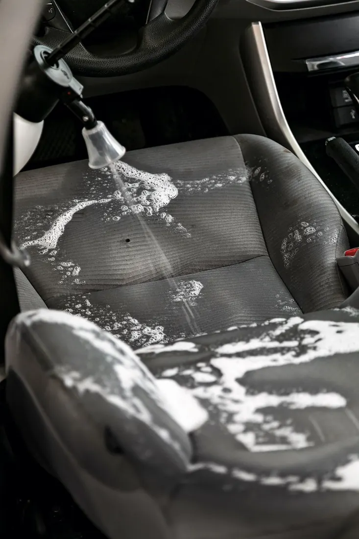 Lavado de vestiduras de auto: asiento de tela gris recibiendo shampoo en espuma con boquilla de rociado